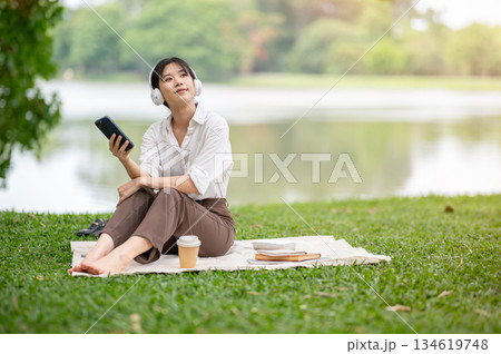 Asian woman wearing headphone listening to music while holding phone sitting at picnic mat on grass. 134619748