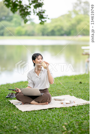 Asian woman sipping or drinking coffee while sitting with laptop on picnic mat laying on the grass. 134619749