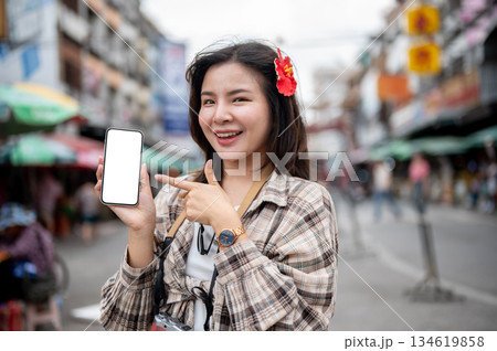 Asian tourist woman with red flower on hair showing and pointing at white screen phone in a market. Asian tourist woman with red flower on hair showing and pointing at white screen phone in a market. 134619858