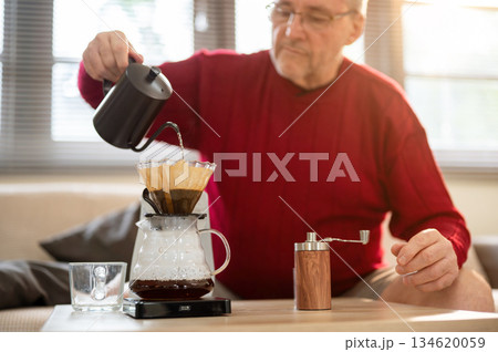 Glasses old white man pouring hot water from kettle into coffee in filter paper on dripper at table. Glasses old white man pouring hot water from kettle into coffee in filter paper on dripper at table. 134620059