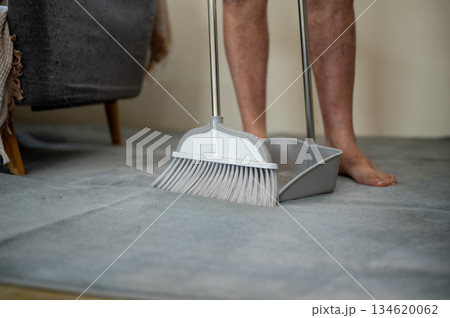 Close up of an old man standing holding a broom sweeping the dust off carpet and floor in his room 134620062