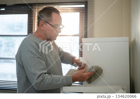 Old white man with glasses spraying a cleaning liquid on cloth rag wiping the dust off in his room. Old white man with glasses spraying a cleaning liquid on cloth rag wiping the dust off in his room. 134620066