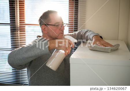 Old white caucasian man holding cleaning foggy spray and cloth rag wiping dust off shelf in his room Old white caucasian man holding cleaning foggy spray and cloth rag wiping dust off shelf in his room 134620067