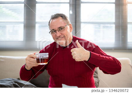 Old white caucasian man wearing glasses holding his coffee and making a thumb up on living room sofa 134620072