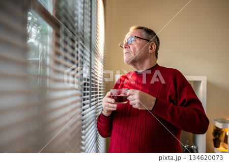 Old white caucasian man wearing glasses holding his coffee cup looking ahead through window blinds. Old white caucasian man wearing glasses holding his coffee cup looking ahead through window blinds. 134620073
