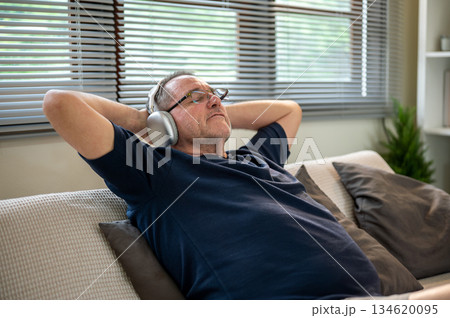 Old white caucasian man with glasses and headphones with hands behind head leaning on sofa in house. Old white caucasian man with glasses and headphones with hands behind head leaning on sofa in house. 134620095