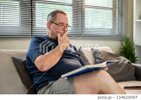Old white caucasian man wearing glasses looking curious as reading a book sitting on sofa in house. 134620097