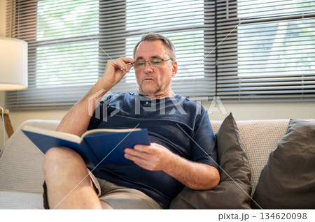 Old white caucasian man touching his glasses as holding book reading while sitting on sofa in house. 134620098