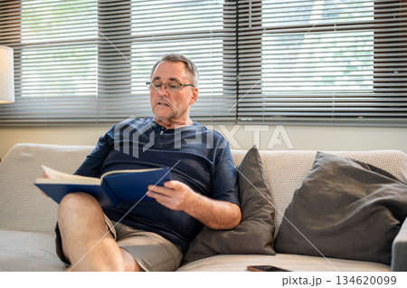 Old white caucasian man wearing glasses holding book reading calmly as sitting on sofa in the house. 134620099