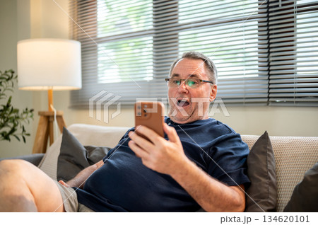 Old white caucasian man wearing glasses looking excited at phone in hand as sitting on sofa in house 134620101