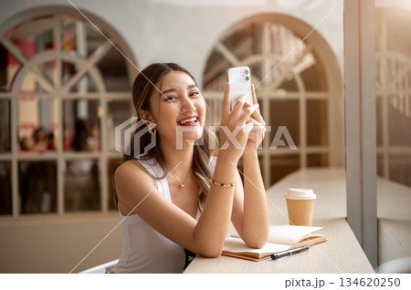 Smiling asian woman looking away from phone holding in hands with notebook and coffee on wooden table 134620250
