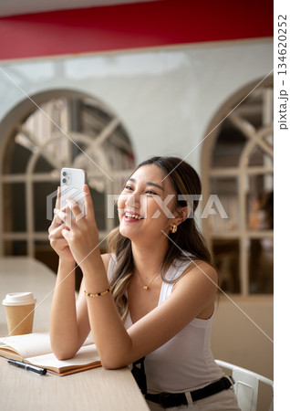 Asian woman laughing as looking at phone hold in both hands with notebook and coffee on wooden table Asian woman laughing as looking at phone hold in both hands with notebook and coffee on wooden table 134620252
