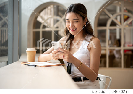 Asian woman smiling while looking at phone holds in hands with notebook and coffee on wooden table. 134620253