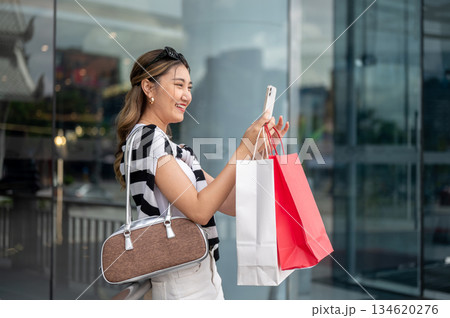 Asian woman holding shopping bags taking photos on phone standing aside railing in front of a mall. Asian woman holding shopping bags taking photos on phone standing aside railing in front of a mall. 134620276