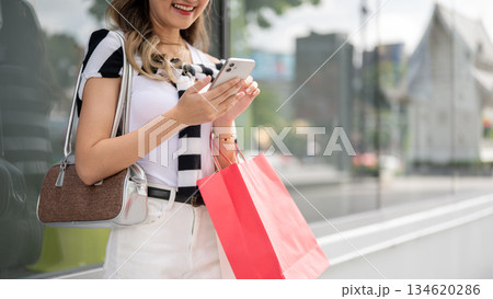 Smiling woman holding shopping bags looking at phone standing aside glass display cabinet at mall. Smiling woman holding shopping bags looking at phone standing aside glass display cabinet at mall. 134620286