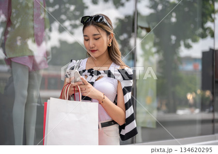 Asian woman holding shopping bag texting on phone near mannequin in the mall's glass display cabinet Asian woman holding shopping bag texting on phone near mannequin in the mall's glass display cabinet 134620295