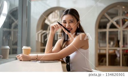 Asian woman in tank top holding pen in front of her face with phone and coffee aside on wooden table Asian woman in tank top holding pen in front of her face with phone and coffee aside on wooden table 134620301