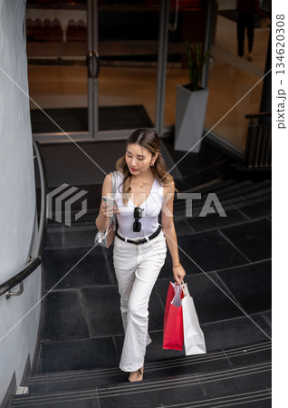 Asian woman in tank top holds shopping bags looking at phone walking upstair from mall underground. Asian woman in tank top holds shopping bags looking at phone walking upstair from mall underground. 134620308