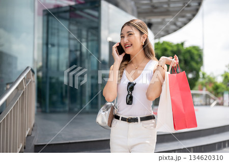 Smiling asian woman in white tank top holding shopping bags during a phone call in the mall's forecourt Smiling asian woman in white tank top holding shopping bags during a phone call in the mall's forecourt 134620310
