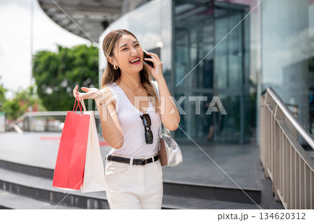 Smiling asian woman in white tank top holding shopping bags talking on phone in the mall's forecourt Smiling asian woman in white tank top holding shopping bags talking on phone in the mall's forecourt 134620312