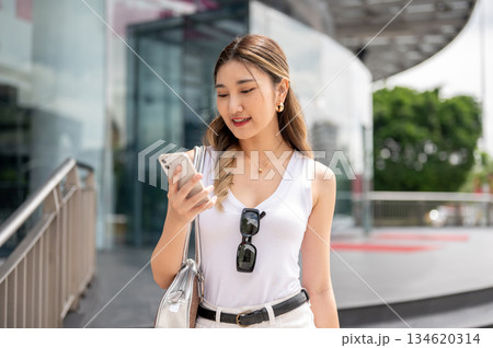 Smiling asian woman in white tank top holding and looking at phone while standing in mall forecourt 134620314