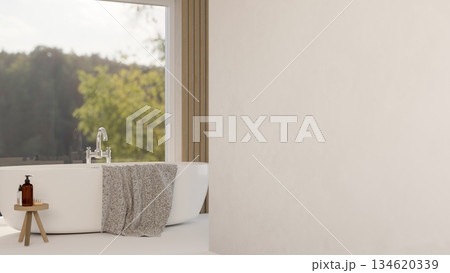 Ceramic bathtub with body wash atop wooden stool chair on white floor in a bathroom with glass wall. 134620339