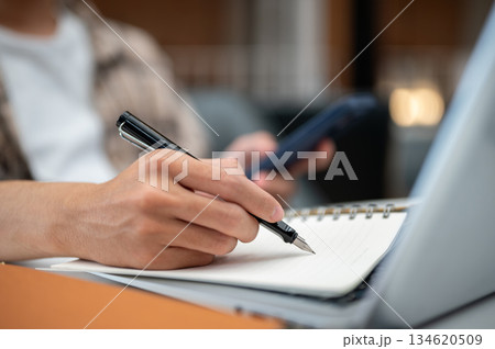 Close up of a man hands holding phone and writing in notebook with a pen over sits at table in cafe. Close up of a man hands holding phone and writing in notebook with a pen over sits at table in cafe. 134620509