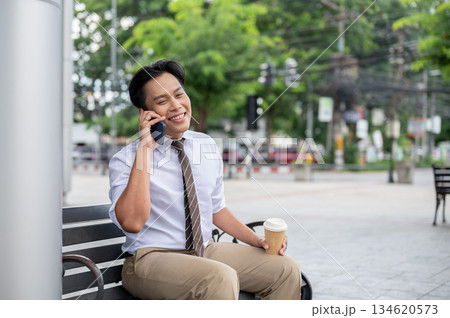 Male office worker holding coffee cup and talking happily on phone while sitting on steel bench in the park. 134620573