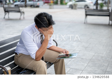 Male office worker massaging or rubbing temple while holding tablet sitting on steel bench in park. 134620588
