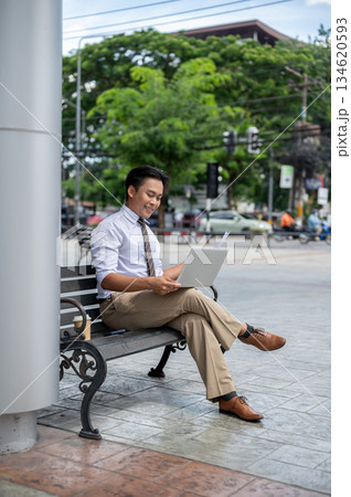 Smiling male office worker looking at his laptop while sitting cross legged on steel bench in park. 134620593