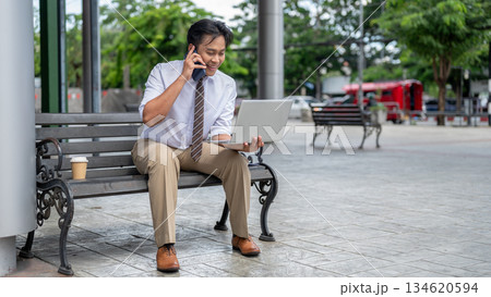 Smiling male office worker talking on phone looking at laptop while sitting on steel bench in park. 134620594