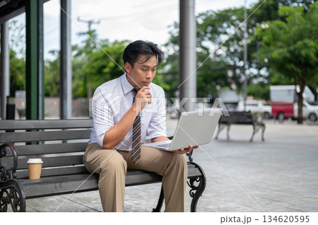 Male office worker holding and looking at laptop with hand under chin while sitting on bench in park 134620595