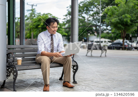 Smiling male office worker looking at laptop holding with both hands while sitting on bench in park. 134620596