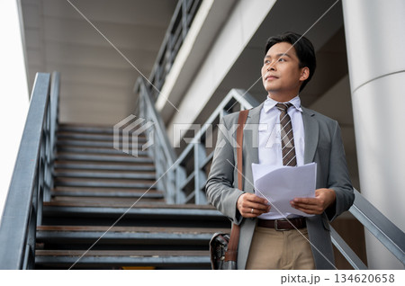 Businessman or office worker looking away from documents and standing on flyover or overpass stairs. Businessman or office worker looking away from documents and standing on flyover or overpass stairs. 134620658