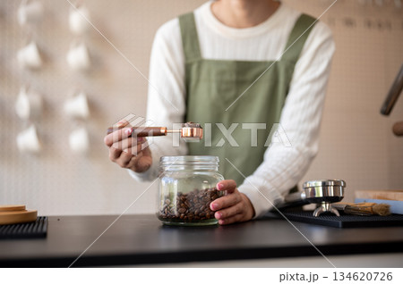 Close up of a green apron barista holding spoon scooping coffee beans from glass jar at counter bar. 134620726