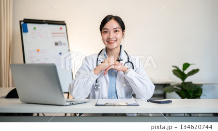 Asian female doctor holding pen with both hands under chin with a laptop on table in diagnosis room. 134620744