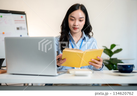 Female office worker or businesswoman reading yellow cover book with laptop aside on working table. Female office worker or businesswoman reading yellow cover book with laptop aside on working table. 134620762