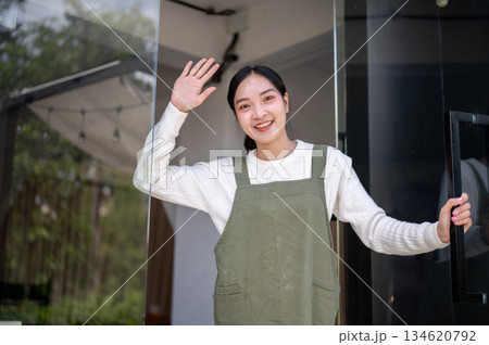 Asian female barista or owner in green apron holding or opening glass door waving hand for customers 134620792