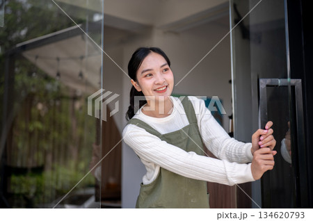 Asian female barista or owner in green apron holding or opening cafe glass door waiting for customer Asian female barista or owner in green apron holding or opening cafe glass door waiting for customer 134620793
