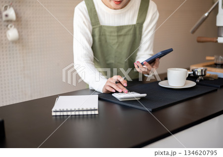 Close up of female barista or owner in green apron holding phone using a calculator on counter bar. 134620795