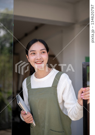Asian female barista in green apron holding notebook and opening a glass door waiting for customers. 134620797