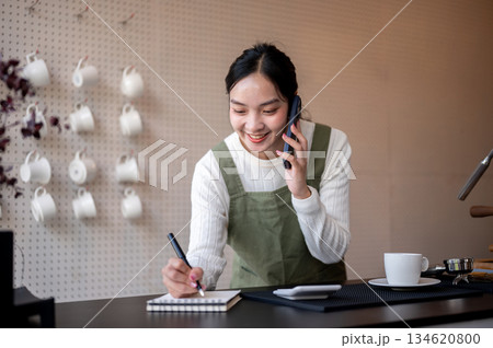 Female barista or owner in green apron talking on phone writing in notebook with pen on counter bar. 134620800