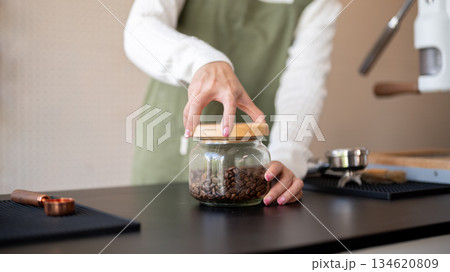 Close up of green apron barista hand opening or closing lid of coffee beans jar at cafe counter bar. Close up of green apron barista hand opening or closing lid of coffee beans jar at cafe counter bar. 134620809