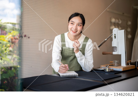Asian female barista in green apron holding pencil thinking new menu in notebook at cafe counter bar Asian female barista in green apron holding pencil thinking new menu in notebook at cafe counter bar 134620821