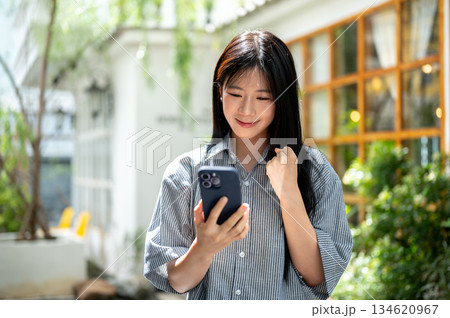 Smiling asian woman raising a fist and looking at smartphone while standing in cafe's outdoor garden 134620967