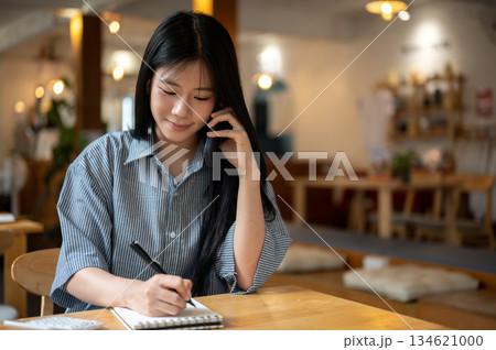 Asian woman talking on phone holding pen writing in notebook with calculator on wooden table in cafe 134621000