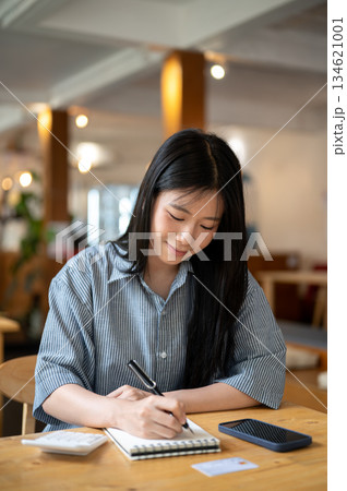 Asian woman smiling while holding pen writing in notebook with calculator and phone on cafe's table. 134621001