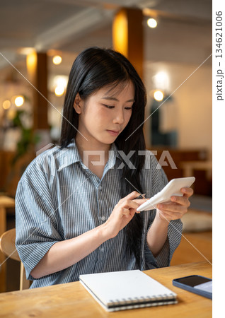Asian woman holding and using calculator with notebook and phone aside sits at wooden table in cafe. Asian woman holding and using calculator with notebook and phone aside sits at wooden table in cafe. 134621006