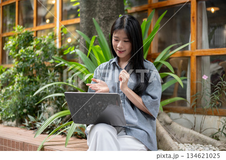 Asian woman explaining with hand while sitting cross legged looking at laptop at cafe outdoor garden 134621025
