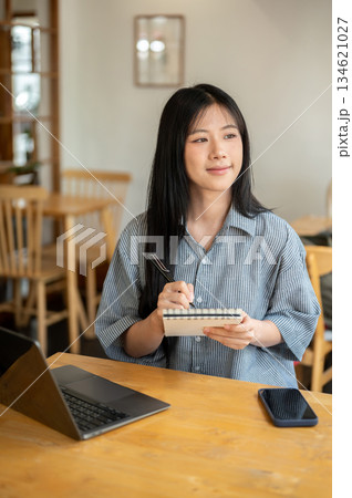Smiling asian woman holding pen looking away from notebook with laptop aside on wooden table in cafe Smiling asian woman holding pen looking away from notebook with laptop aside on wooden table in cafe 134621027